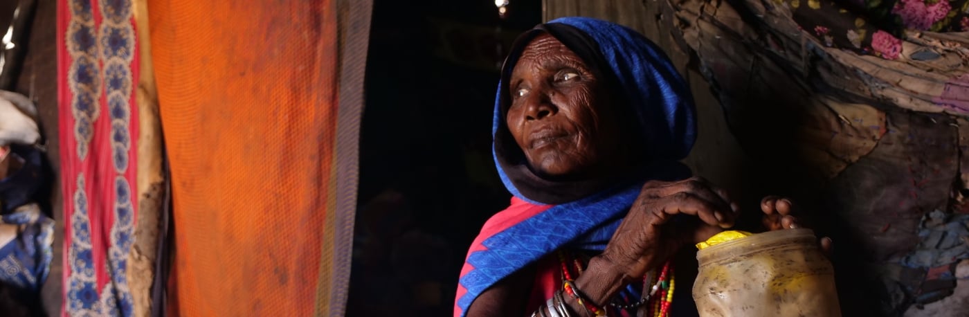 An older woman sitting inside a traditional home, holding a container, and dressed in vibrant tribal attire with beadwork.